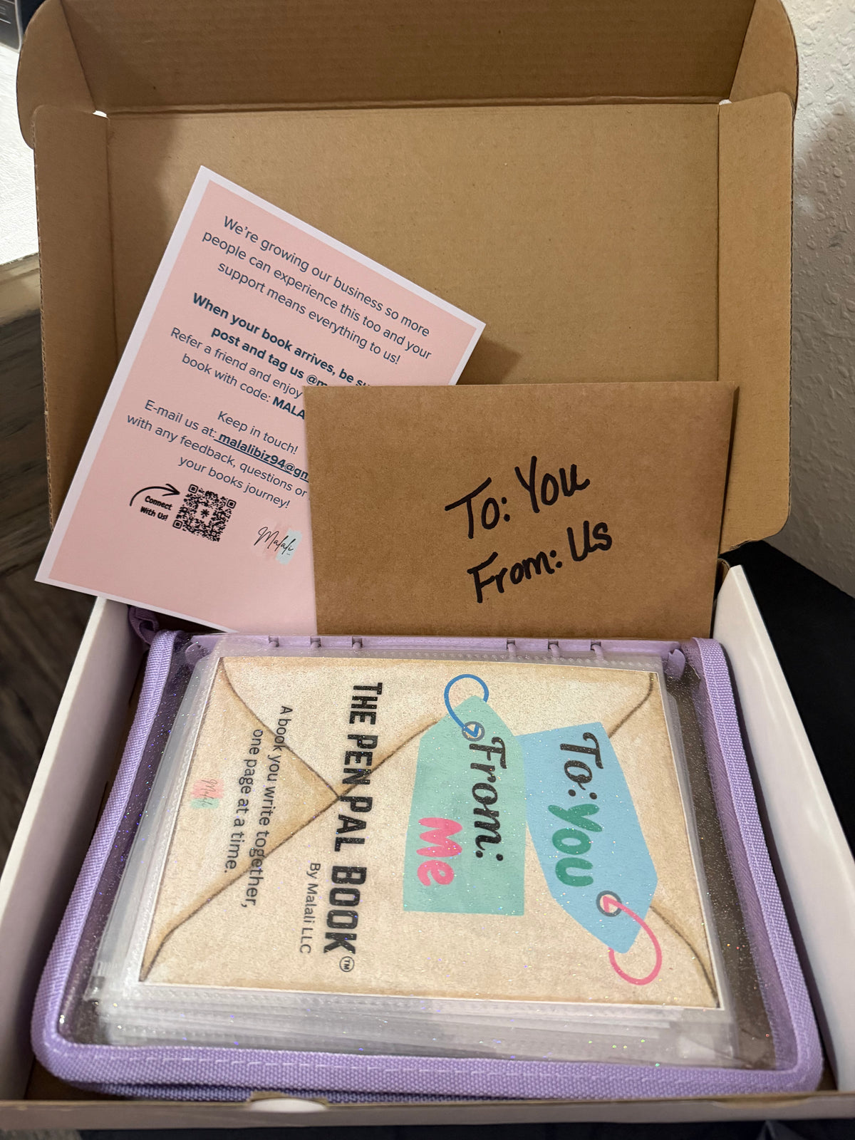 A boxed book package shown open, with a kraft paper insert reading 'To: You From: Us' and a small card. Inside the box is a book titled 'The Penpal Book' in a clear sleeve with a purple border and a matching tag design.