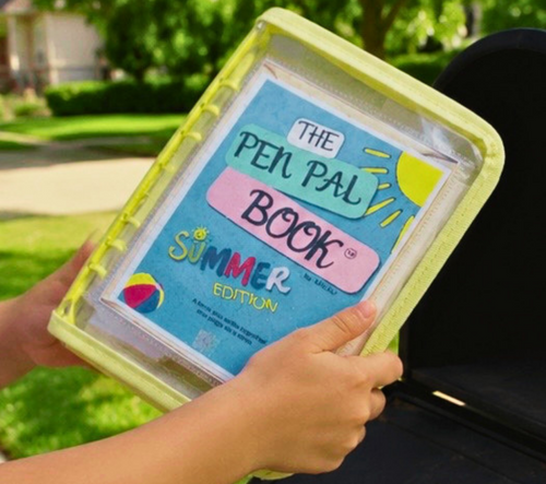Person holding 'The Pen Pal Book Summer Edition' in a clear plastic case outdoors.